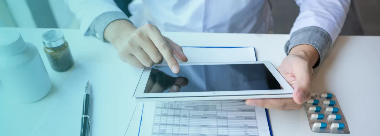طبيب يجلس على مكتبه ويطالع تابلت وعلى المكتب تقرير تحليل طبي فيه اختصارات التحاليل